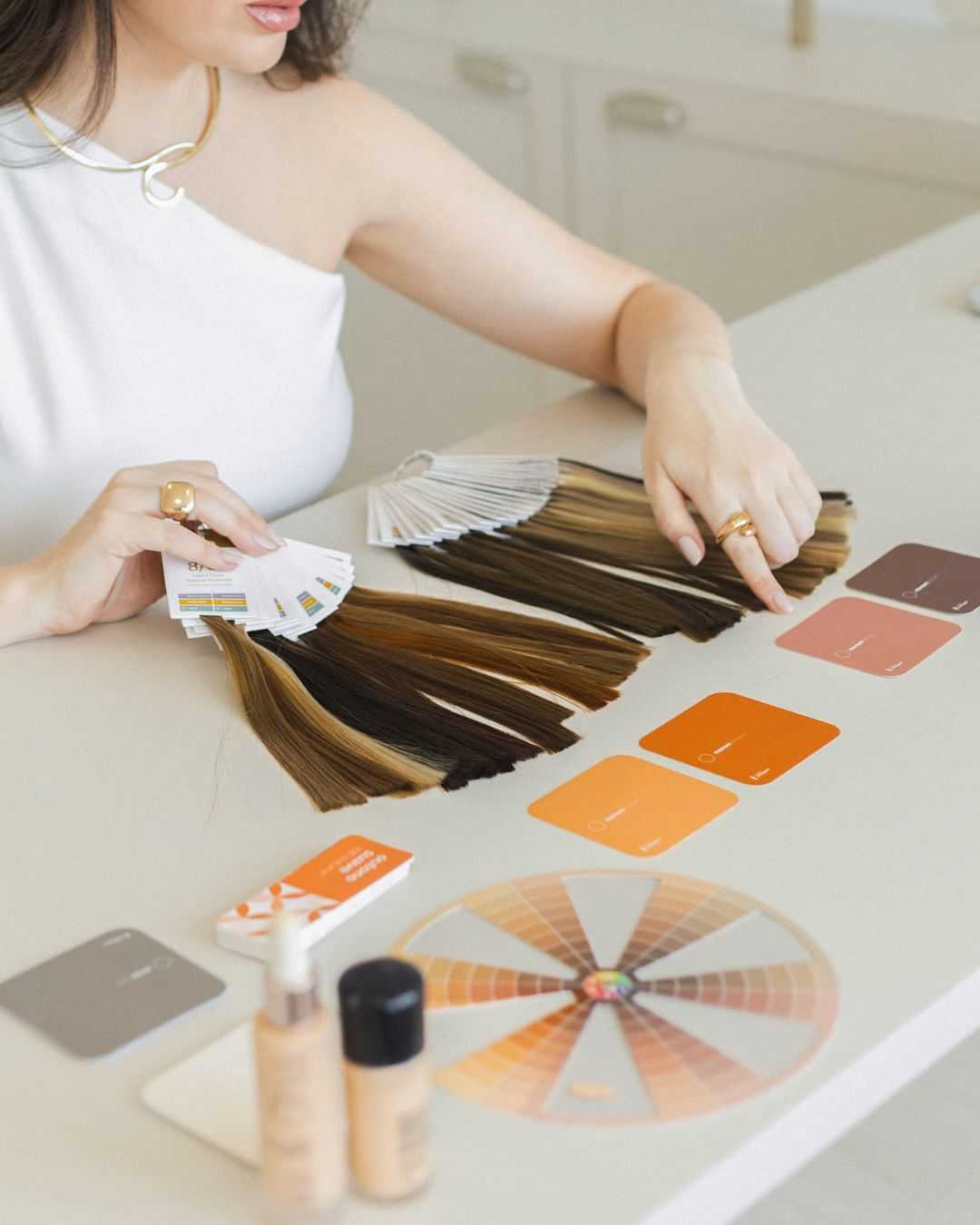 Woman selecting hair color samples from a palette, with makeup and color charts on a table.