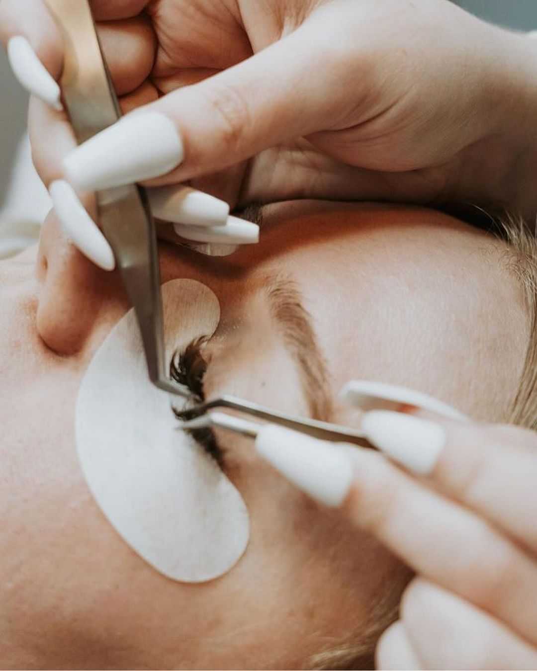 Close-up of person receiving eyelash extension treatment with tweezers and eye patch.